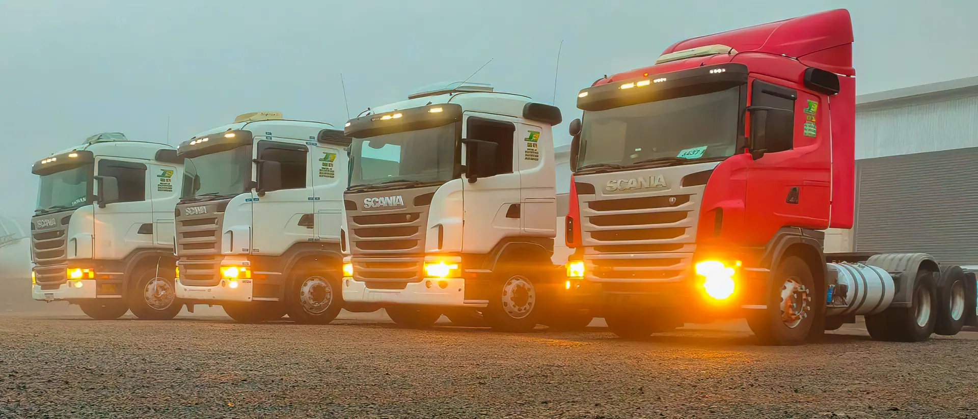 Quatro caminhões da J3 Transportes lado a lado com os faróis acesos em uma manhã de neblina